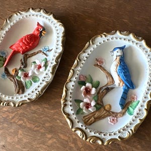 May include: Two oval ceramic wall plaques with gold-coloured ornate trim. One shows a red cardinal on a branch with flowers. The other depicts a blue jay on a branch with flowers.