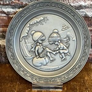 Peut inclure: Une assiette en étain argentée avec un motif en relief de deux enfants faisant du patin à glace dans une scène d'hiver. L'assiette a une bordure de feuilles de houx et le texte "Joan Walsh Anglund" en bas.