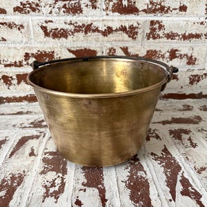 May include: A vintage brass bucket with a black handle. The bucket has a rounded shape and a shiny, aged finish. The background is a white brick wall with brown accents.