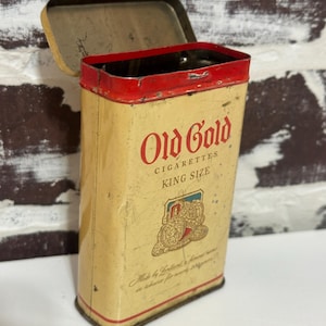Könnte beinhalten: Vintage "Old Gold" King Size Zigarettendose. Die rechteckige Dose hat einen cremefarbenen Hintergrund mit roten und goldenen Akzenten. Der Deckel ist geöffnet und gibt den Blick ins Innere frei. Der Text "Old Gold Cigarettes" ist rot gedruckt, zusammen mit einer Grafik.