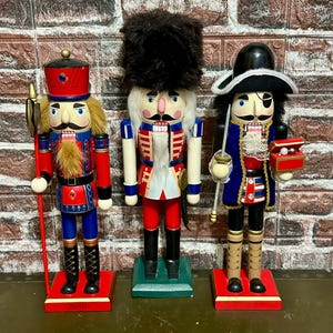 May include: Three decorative wooden nutcrackers in military-style uniforms. The figures are painted in red, blue, and black with gold detailing. One holds a spear, another a sword, and the third a small box. Each stands on a base.