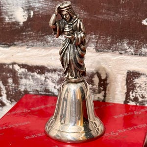 May include: A silver bell with a detailed figurine of a person on top. The figurine is wearing flowing robes and holding an object. The bell is on a red box with the words "REED & BARTON" printed on it.