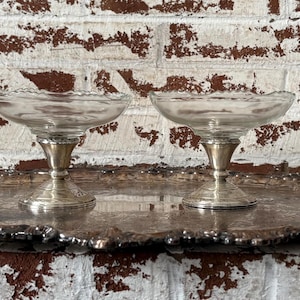 May include: Two clear glass dessert bowls with scalloped edges sit on silver-colored pedestals. They are displayed on a silver-colored, ornate tray. The background is a white brick wall with brown paint.