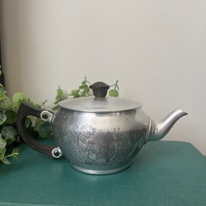 Puede incluir: Una tetera de plata con asa y pomo negros. La tetera tiene un diseño grabado de árboles y una pagoda.