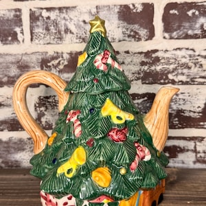 Puede incluir: Una tetera de cerámica con forma de árbol de Navidad y una estrella dorada en la parte superior. El árbol verde está decorado con bastones de caramelo, adornos y cuernos amarillos. La base de la tetera tiene un diseño de caja de regalo y tambor.