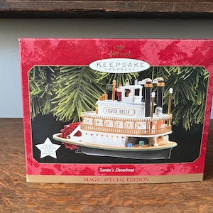Könnte beinhalten: Hallmark Keepsake-Ornament in einer roten Box. Das Ornament ist ein Miniatur-Raddampfer namens "Silver Belle". Auf der Box steht "Santa's Showboat" und "Magic Special Edition". Das Ornament hat Licht- und Bewegungsfunktionen.