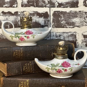 Könnte beinhalten: Zwei weiße Porzellan-Öllampen mit einem floralen Rosendesign. Jede Lampe hat einen Messingbrenner und einen Griff. Die Lampen stehen auf antiken Büchern mit dem Titel "The Class Room Teacher".