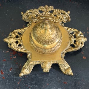 May include: A gold-colored metal inkwell featuring an ornate, symmetrical base with intricate scrollwork and floral detailing. The central component is a domed, circular lid with engraved patterns, resting on a square platform with four decorative feet.