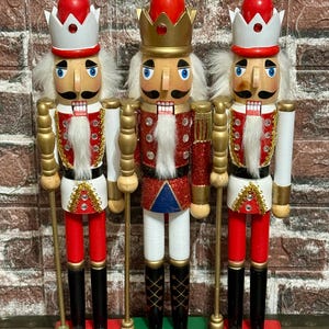 May include: Three wooden nutcrackers in festive attire. Each figure has a red and white crown, a white beard, and a red jacket with gold accents. They stand on red and green bases, holding gold staffs. A classic holiday decoration.