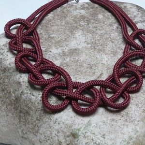 May include: A burgundy-colored necklace with a knotted design. The necklace is made of thick, woven material and features a series of interconnected loops. The necklace is displayed on a textured, gray stone surface.
