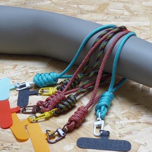 May include: A collection of colorful phone lanyards with various rope designs and metal clasps. The lanyards feature a variety of colors, including teal, red, yellow, and black. The lanyards are attached to a gray cylindrical object.