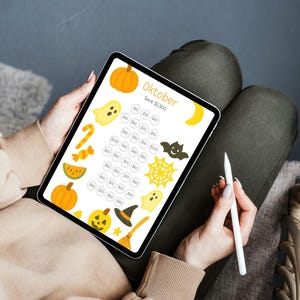 May include: A digital Halloween-themed savings tracker on a tablet. The screen displays the word "Oktober" and various Halloween icons like pumpkins, ghosts, and bats. A stylus is held near the tablet.