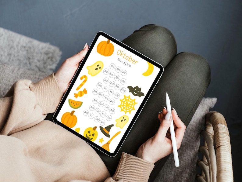 May include: A digital Halloween-themed savings tracker on a tablet. The screen displays the word "Oktober" and various Halloween icons like pumpkins, ghosts, and bats. A stylus is held near the tablet.