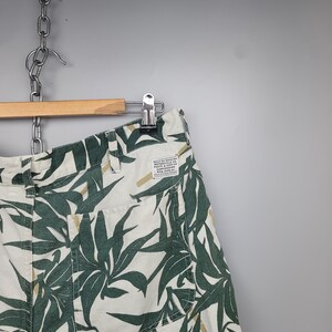 May include: Off-white shorts featuring a dark green leaf pattern and tan accents. The shorts include a back pocket and a rectangular white label with the text "DEUS EX MACHINA MOTORCYCLE CO. PARTE & SERVICE CAMPERDOWN NSW 2050 AU".