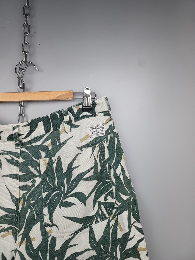 May include: Off-white shorts featuring a dark green leaf pattern and tan accents. The shorts include a back pocket and a rectangular white label with the text "DEUS EX MACHINA MOTORCYCLE CO. PARTE & SERVICE CAMPERDOWN NSW 2050 AU".