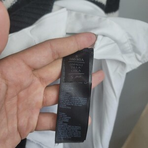 Pu&ograve; includere: Un'etichetta nera con testo bianco, tenuta contro una camicia bianca e un maglione a righe bianche e nere. L'etichetta recita "S 160/80A (SMALL) TALLA: CHICA" e include le istruzioni di lavaggio. Il maglione ha una trama a coste.