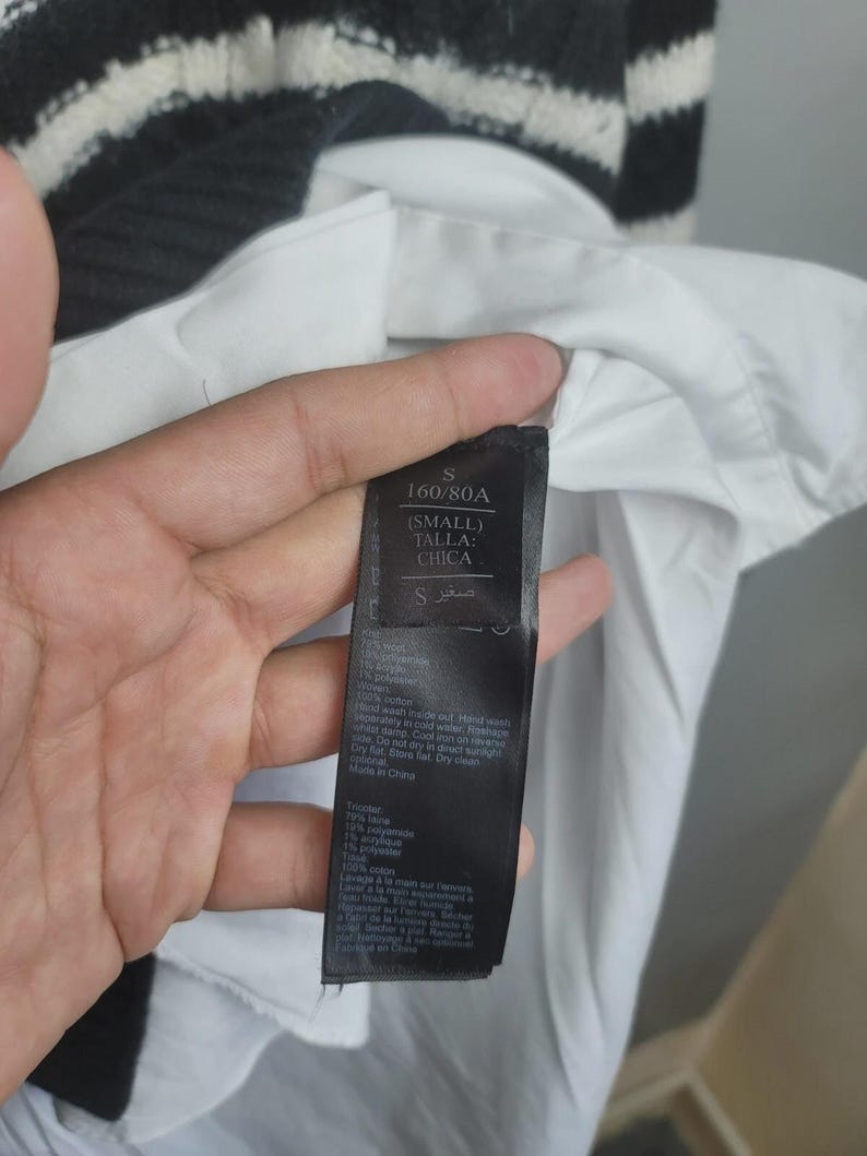 Pu&ograve; includere: Un'etichetta nera con testo bianco, tenuta contro una camicia bianca e un maglione a righe bianche e nere. L'etichetta recita "S 160/80A (SMALL) TALLA: CHICA" e include le istruzioni di lavaggio. Il maglione ha una trama a coste.