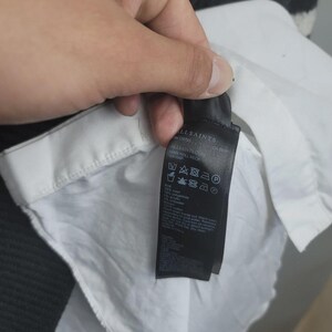 Pu&ograve; includere: Un'etichetta di abbigliamento nera con testo bianco &egrave; tenuta contro una camicia bianca. L'etichetta mostra il marchio "ALLSAINTS". La camicia ha un colletto bianco e si vede un maglione a righe bianche e nere.