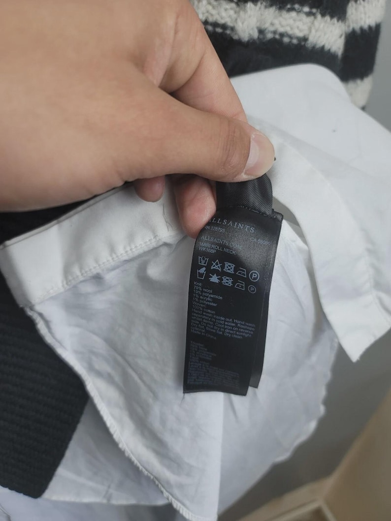 Pu&ograve; includere: Un'etichetta di abbigliamento nera con testo bianco &egrave; tenuta contro una camicia bianca. L'etichetta mostra il marchio "ALLSAINTS". La camicia ha un colletto bianco e si vede un maglione a righe bianche e nere.