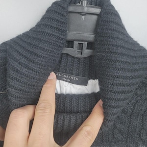 Pu&ograve; includere: Un maglione di maglia grigio scuro con collo ampio e strisce bianche sul fondo. L'etichetta recita "ALLSAINTS SMALL". Il maglione &egrave; appeso a una gruccia nera.