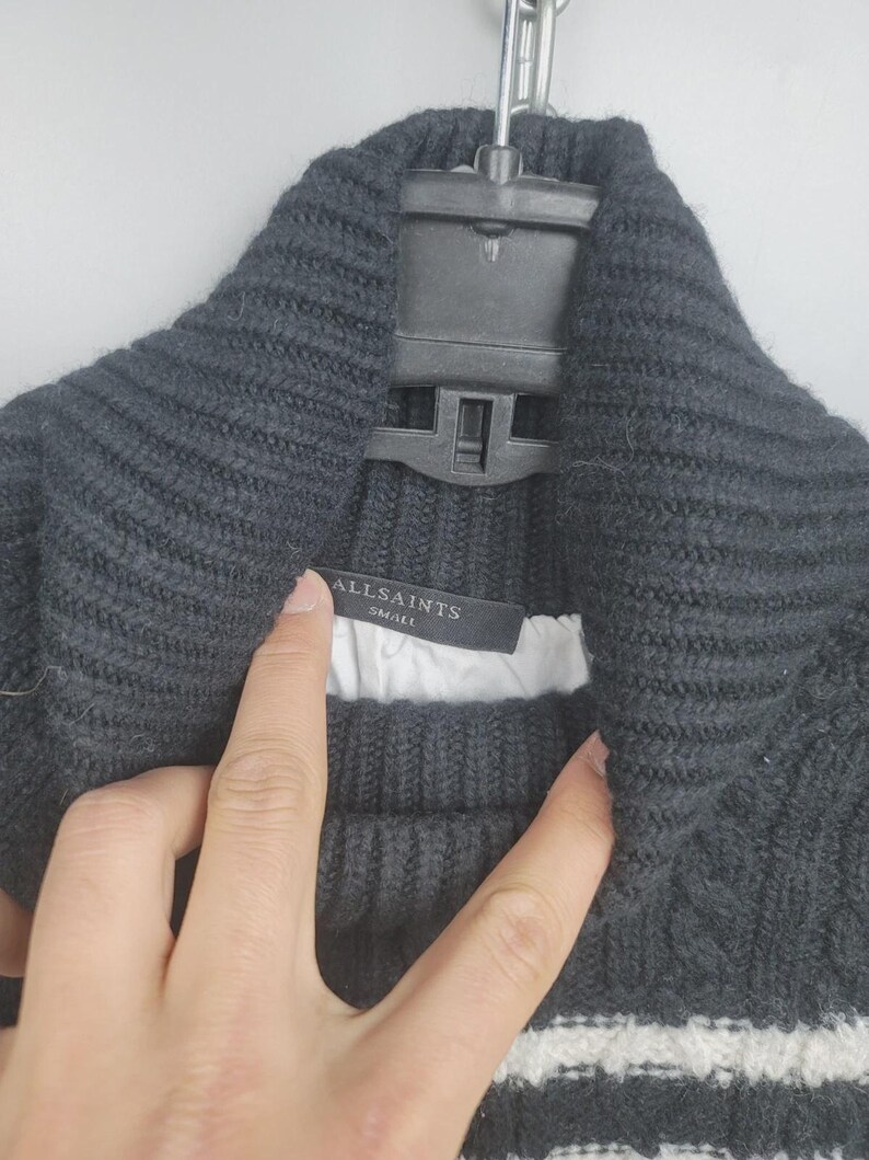 Pu&ograve; includere: Un maglione di maglia grigio scuro con collo ampio e strisce bianche sul fondo. L'etichetta recita "ALLSAINTS SMALL". Il maglione &egrave; appeso a una gruccia nera.