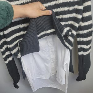 Pu&ograve; includere: Un maglione a righe grigie e bianche con una camicia bianca sotto. Il maglione ha maniche lunghe e un orlo a coste. La camicia &egrave; visibile nella parte inferiore del maglione.