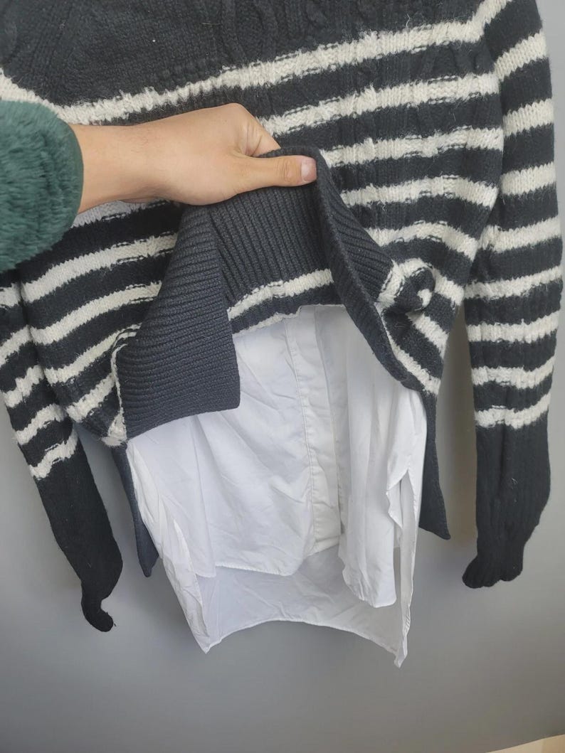Pu&ograve; includere: Un maglione a righe grigie e bianche con una camicia bianca sotto. Il maglione ha maniche lunghe e un orlo a coste. La camicia &egrave; visibile nella parte inferiore del maglione.