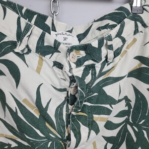 May include: Off-white shorts featuring a dark green leaf and bamboo print. The shorts have a button-fly closure with four visible buttons and a waistband label indicating a 34 inch waist size. The fabric has a visible twill weave texture.