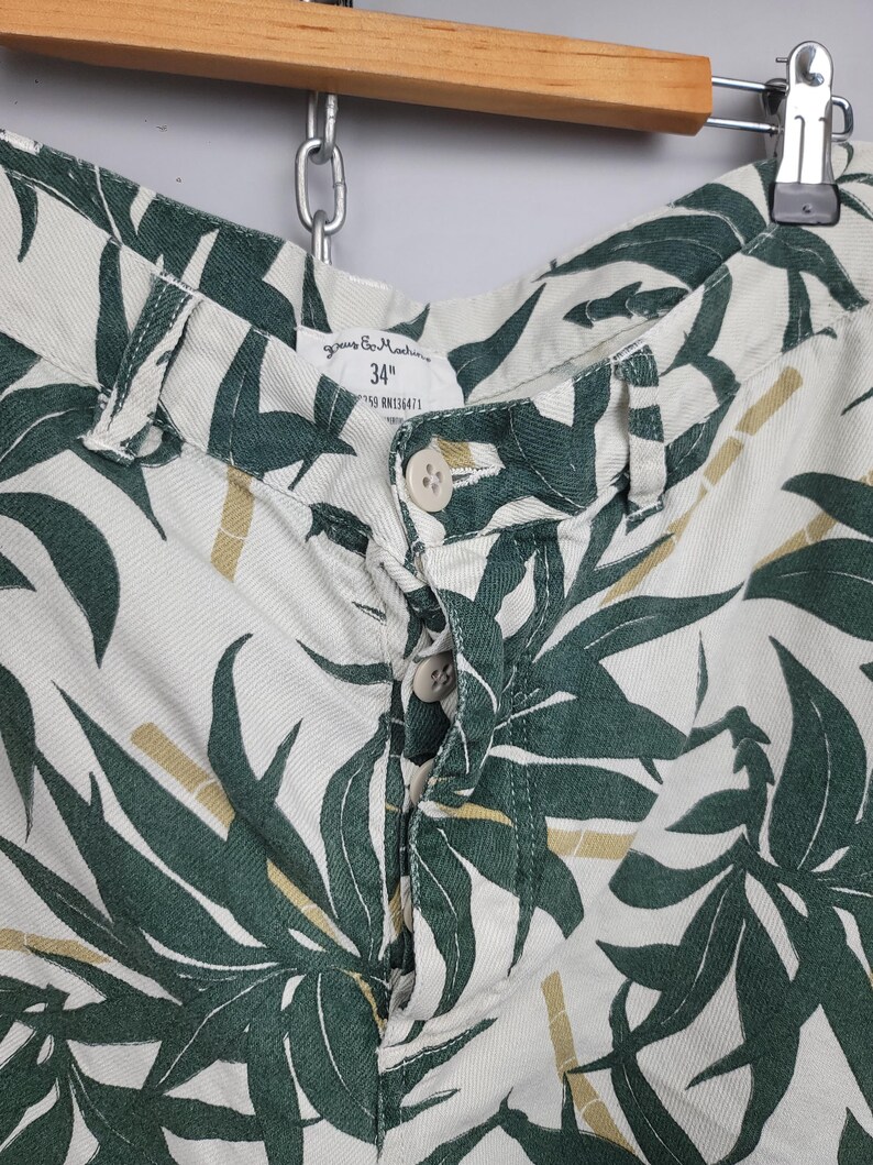 May include: Off-white shorts featuring a dark green leaf and bamboo print. The shorts have a button-fly closure with four visible buttons and a waistband label indicating a 34 inch waist size. The fabric has a visible twill weave texture.