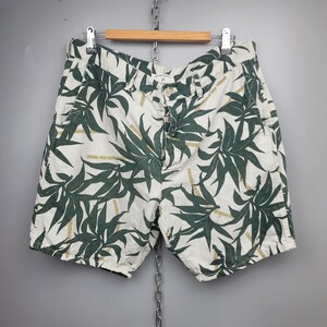May include: Off-white shorts featuring a dark green botanical leaf pattern and light brown bamboo stalk accents. The shorts include a button fly closure, belt loops, and side pockets, presented on a wooden hanger against a gray background.