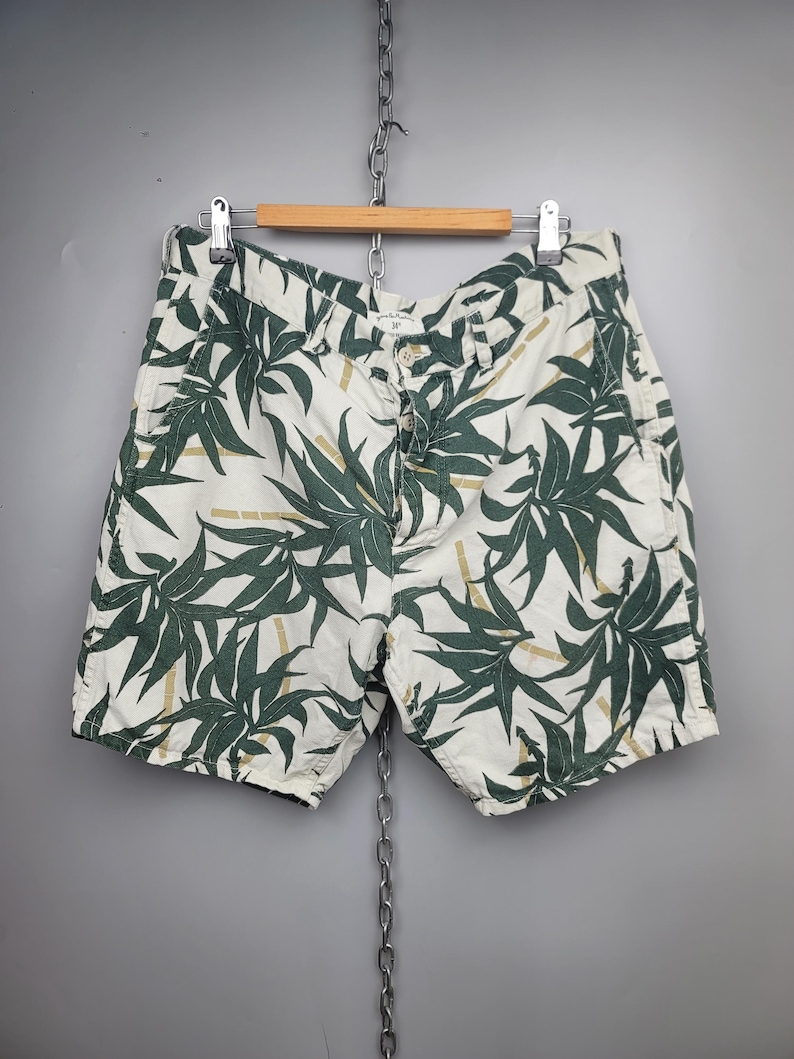 May include: Off-white shorts featuring a dark green botanical leaf pattern and light brown bamboo stalk accents. The shorts include a button fly closure, belt loops, and side pockets, presented on a wooden hanger against a gray background.