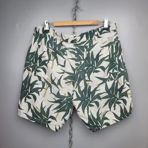 May include: Off-white shorts featuring a dark green botanical leaf and bamboo stalk print. The shorts include two back pockets with button closures, a waistband with belt loops, and a small rectangular white brand label on the upper right side.