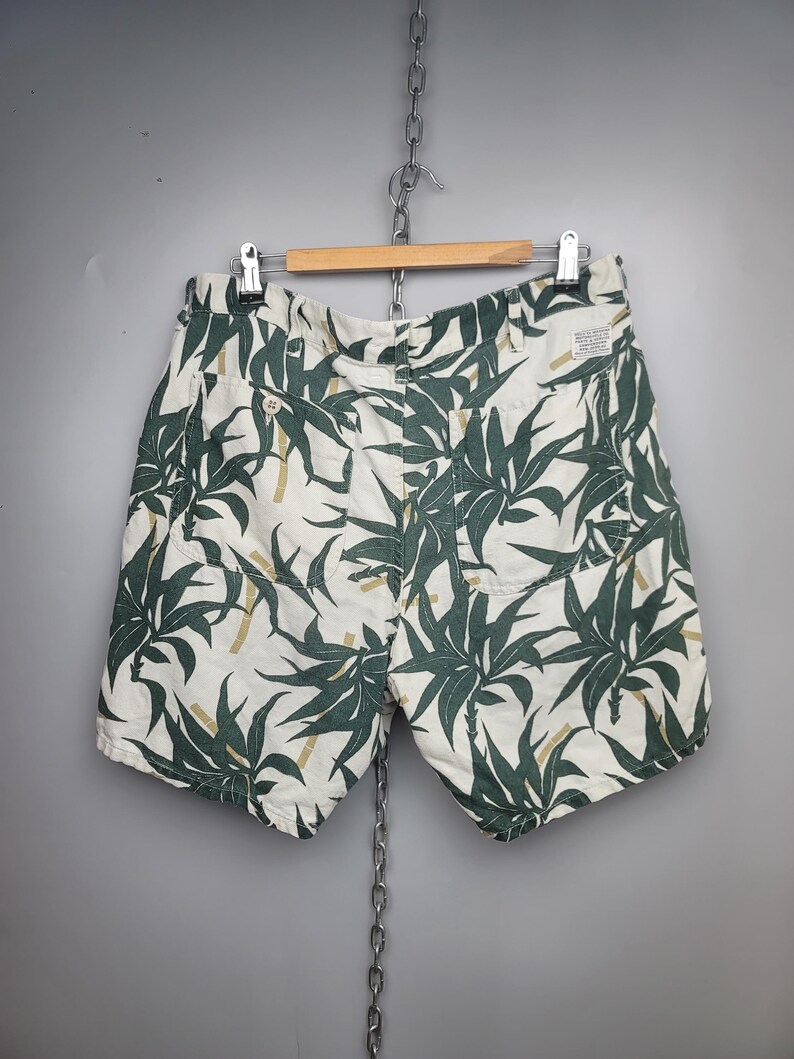 May include: Off-white shorts featuring a dark green botanical leaf and bamboo stalk print. The shorts include two back pockets with button closures, a waistband with belt loops, and a small rectangular white brand label on the upper right side.