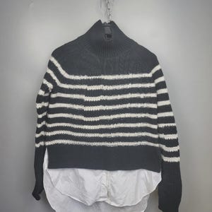 Pu&ograve; includere: Un maglione a righe bianche e nere con collo alto e maniche lunghe. Il maglione ha un orlo a camicia bianco. L'indumento &egrave; appeso a una catena di metallo su uno sfondo grigio.