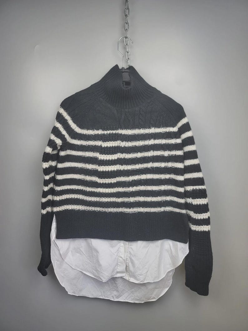 Pu&ograve; includere: Un maglione a righe bianche e nere con collo alto e maniche lunghe. Il maglione ha un orlo a camicia bianco. L'indumento &egrave; appeso a una catena di metallo su uno sfondo grigio.