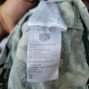 May include: A white fabric garment tag featuring the Deus Ex Machina Motorcycles logo, company address in Camperdown, NSW, Australia, and contact information for an authorized distributor in Seoul, South Korea, against a light green patterned fabric.