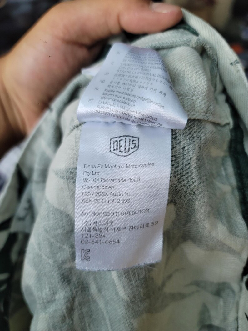 May include: A white fabric garment tag featuring the Deus Ex Machina Motorcycles logo, company address in Camperdown, NSW, Australia, and contact information for an authorized distributor in Seoul, South Korea, against a light green patterned fabric.