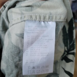 May include: A white fabric garment with a dark green leaf pattern featuring a sewn-in care label. The label displays the text BAMBOO CAMP SHORT, MADE IN CHINA, and lists a material composition of 70% cotton and 30% linen, along with laundry symbols.