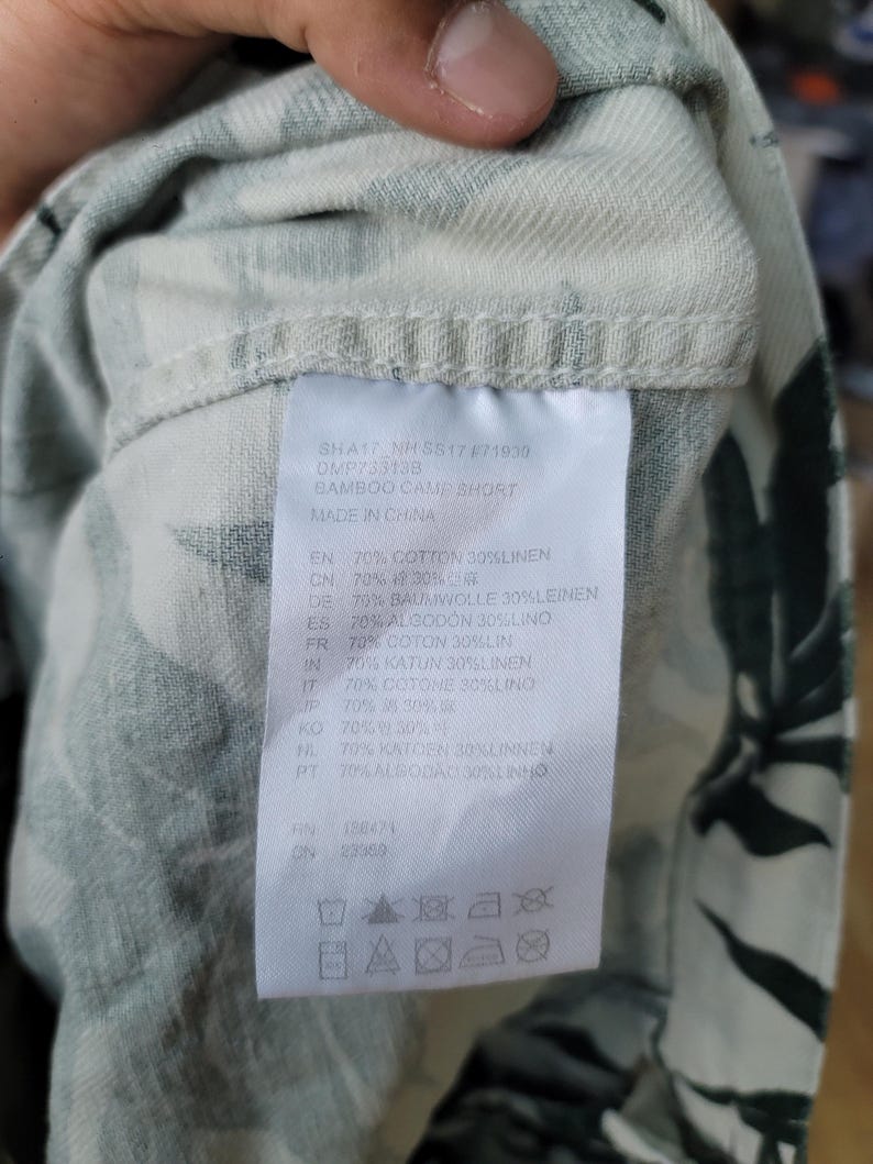 May include: A white fabric garment with a dark green leaf pattern featuring a sewn-in care label. The label displays the text BAMBOO CAMP SHORT, MADE IN CHINA, and lists a material composition of 70% cotton and 30% linen, along with laundry symbols.