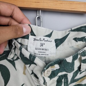 May include: Off-white shorts featuring a dark green leaf pattern. The interior label displays the brand name Deus Ex Machina, a 34 inch waist size, CA23359 RN136471, the phrase In Benzin Veritas, and Made in China. A single beige button is visible.