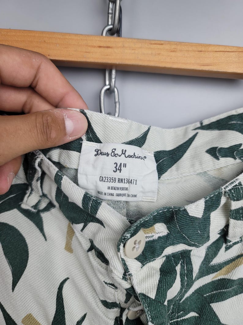 May include: Off-white shorts featuring a dark green leaf pattern. The interior label displays the brand name Deus Ex Machina, a 34 inch waist size, CA23359 RN136471, the phrase In Benzin Veritas, and Made in China. A single beige button is visible.