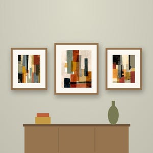 May include: A set of three framed abstract art prints featuring torn paper shapes in shades of rust, olive green, black, and cream. The prints are displayed above a brown wooden sideboard with a small olive vase and stacked boxes.