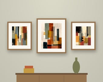May include: A set of three framed abstract art prints featuring torn paper shapes in shades of rust, olive green, black, and cream. The prints are displayed above a brown wooden sideboard with a small olive vase and stacked boxes.