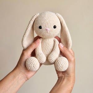 May include: A handmade, cream-coloured crochet bunny with long ears, black eyes, and a pink nose. The stuffed animal is held in two hands, showcasing its soft texture and detailed craftsmanship. A charming, cuddly toy.