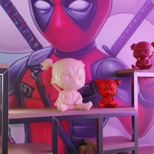 Puede incluir: Tres pequeñas figuras de Deadpool, una en blanco, una en rojo y otra en rojo con una máscara negra. Las figuras están sentadas en un estante de madera con un marco de metal.
