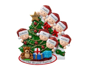 May include: A white Christmas tree ornament with a green Christmas tree, a teddy bear, and presents. Seven children are peeking out from behind the tree, each with their name written on their hat. The text "The Bryant Family" is written at the bottom of the ornament.