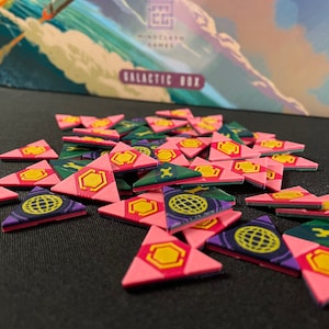 May include: A pile of colourful triangular game pieces with different symbols. The pieces are pink, green, purple, and yellow with a hexagonal, wrench, and globe design. The pieces are from the Galactic Box game.