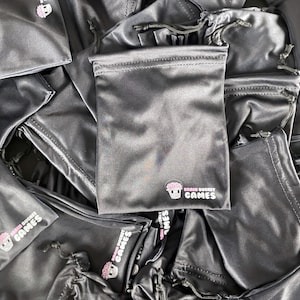 Puede incluir: Un conjunto de bolsas negras de satén con cordón. Cada bolsa tiene un logotipo rosa y blanco que dice "Brain Bucket Games". Algunas bolsas están abiertas, otras cerradas, todas con cierre de cordón.