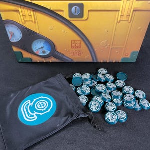 Könnte beinhalten: Eine Spielbox in Gelb und Grün mit einer Messgerätegrafik. Ein schwarzer Kordelzugbeutel mit einem türkisfarbenen Logo schüttet türkisfarbene Spielsteine aus. Die Spielsteine haben ein weißes Spiraldesign.