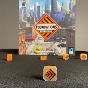 Puede incluir: Primer plano de una pieza de juego para el juego de mesa "Foundations: Metropolis". La pieza es naranja y gris con un diseño de flecha negra. La caja del juego está en el fondo.